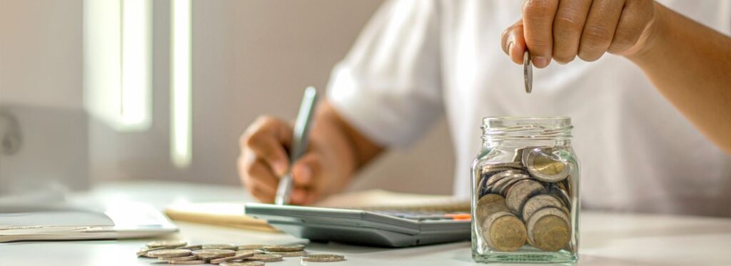 Uma mão a deixar cair uma moeda num frasco cheio de moedas. Mais moedas pousadas numa mesa e uma calculadora, pessoa a fazer contas para poupar com a frota. Reduzir despesa de combustível